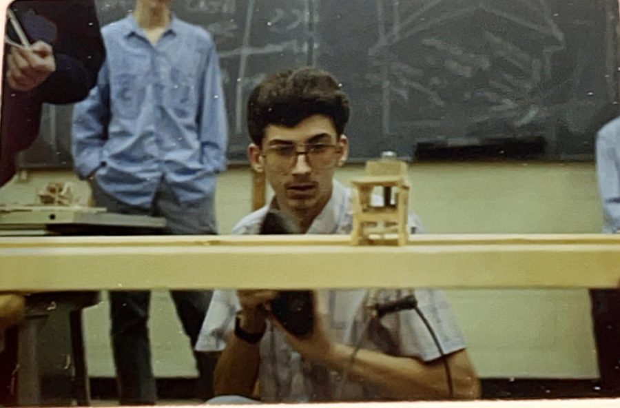 A student gets down low and looks at his bridge design while weight is addedthe blackboards in te back look as though the have the illustration of the days procedure.